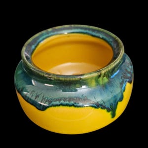 May include: A small, round ceramic bowl with a bright yellow interior and exterior. The rim and upper portion of the bowl are glazed with a blend of green and blue hues, creating a striking contrast. The bowl sits against a black background.