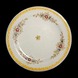 May include: A white ceramic plate with a yellow border and floral design. The plate features a repeating pattern of yellow dashes around the rim and a delicate arrangement of flowers and leaves in the center.