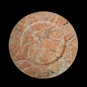 May include: A round plate featuring a natural stone pattern with a mix of salmon, peach, and light brown hues. The surface displays intricate veining in shades of white, grey, and dark charcoal, creating a marbled texture across the entire piece.