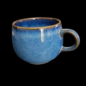 May include: A ceramic mug with a rounded body and a single handle. The exterior features a mottled blue glaze with a darker, brownish-gold rim and handle edge. The interior of the mug is a deep, solid blue color, creating a contrast with the rim.