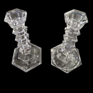 May include: Two clear, faceted crystal candle holders. Each has a hexagonal base, a geometric stem, and a circular top designed to hold a candle. The candle holders are displayed against a black background.