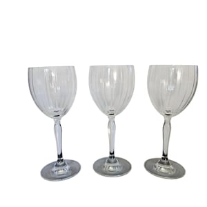 May include: Three clear wine glasses with a ribbed design on the bowl. The glasses have a long, slender stem and a circular base. The glasses are made of clear glass and are set against a white background.