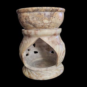 May include: A beige and brown marbled stone oil burner featuring a wide bowl top and a hollow base with carved leaf-like patterns and cut-out details. The natural stone texture displays intricate veining in shades of tan, cream, and dark brown.