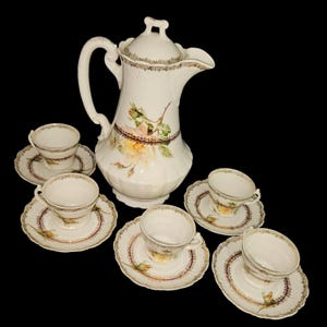 May include: A white porcelain tea set featuring a large coffee pot with a lid and six teacups with saucers. The set is decorated with floral designs in yellow, pink, and green, with gold trim. The background is black.