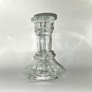 May include: A clear glass candlestick holder with a hexagonal base and a detailed, column-like stem. The top is designed to hold a single candle. The glass is transparent, allowing light to pass through, and the design is classic.