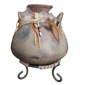 May include: A round ceramic vase with a mottled green, blue, and purple finish. The neck is adorned with brown fur, feathers, and leather strips featuring wooden beads. The vase rests on a dark metal stand with three curved legs.