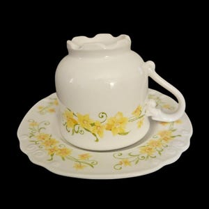 May include: White ceramic teacup and saucer set. The cup has a unique ruffled rim and a curved handle. Both pieces are decorated with yellow floral patterns and green leaves. The saucer has a scalloped edge.