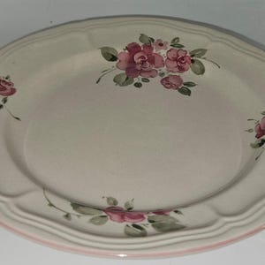 May include: A cream-colored ceramic plate with a scalloped edge and a pink rim. The plate is decorated with pink rose floral patterns and green leaves. A decorative piece for serving food.