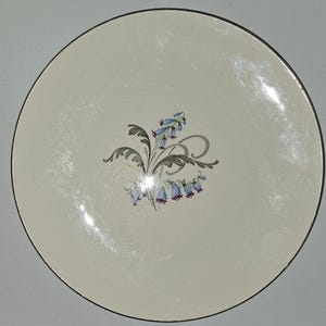 May include: A round, off-white plate with a black rim. The plate features a floral design in the center, with bluebell-like flowers and green leaves. The design is delicate and adds a touch of elegance to the plate.