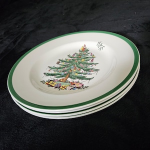 May include: A stack of white ceramic plates featuring a green rim and a central illustration of a decorated Christmas tree with colorful ornaments and wrapped gifts at the base. A small sprig of mistletoe is printed near the upper right edge.