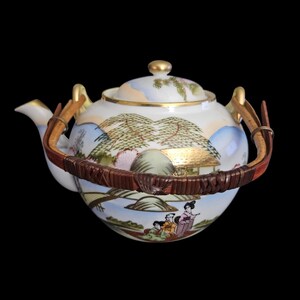 May include: A white ceramic teapot with a landscape design featuring mountains, trees, and figures. The teapot has a gold-colored handle and trim, and a woven brown handle. The lid is topped with a gold knob.