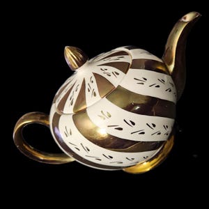 May include: A decorative teapot with a gold and white striped design. The teapot features a curved spout, a rounded handle, and a lid with a gold finial. The design includes small, black, dash-like accents.