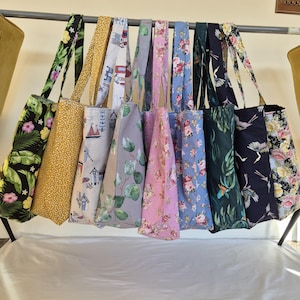 May include: A display of tote bags with diverse floral and patterned designs. The bags are suspended from a metal rail, featuring colours such as black, yellow, blue, pink, and green. Each bag has a coordinating strap.