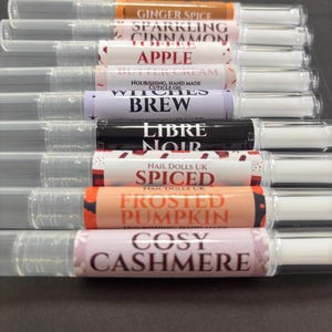 May include: A collection of eight clear plastic cuticle oil pens with white applicator tips. Each pen features a unique label with text including Ginger Spice, Sparkling Cinnamon Toffee, Apple Butter Cream, Witches Brew, Libre Noir, Spiced, Frosted Pumpkin, and Cosy Cashmere.