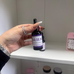 May include: A small dark glass bottle with a black dropper cap and a purple label. The label features the text NAIL DOLLS UK and ALIEN, followed by the words NOURISHING, HAND MADE, FRAGRANCED CUTICLE OIL, and a list of ingredients.