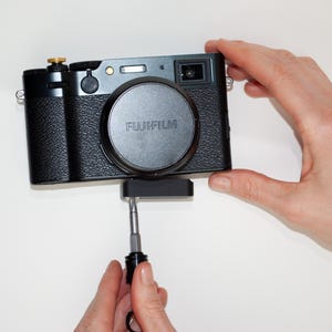 May include: A black Fujifilm digital camera with a lens cap, held by hands. The camera has a textured grip and a silver-colored top button. A small black accessory is attached to the bottom, with a screwdriver being used.
