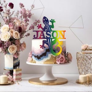 May include: A cake topper with a lizard design and the name "JASON" in rainbow hues. The cake has a marbled effect and is on a white stand. The scene includes flowers and candles, creating a celebratory feel. The cake stand is approximately 20 cm in diameter.