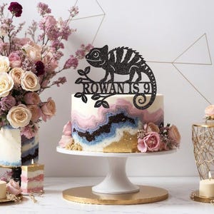 May include: A black glitter chameleon cake topper with the text "Rowan is 9" on a white cake with pink, blue, and gold marbling. The cake is on a white cake stand with a gold base.