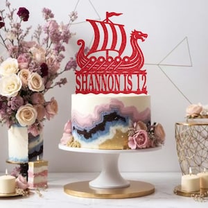 May include: A birthday cake with a red glitter Viking ship cake topper that reads "SHANNON IS 12". The cake has a colourful, marbled design with pink, blue, and gold hues. The cake sits on a white cake stand.