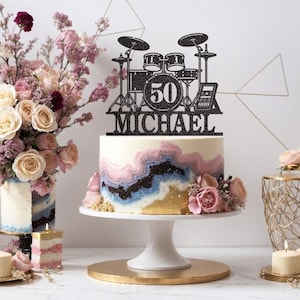 May include: Black glitter cake topper featuring a drum set and the number 50, with the name Michael underneath. The topper is on a white cake with pink, blue, and gold frosting.