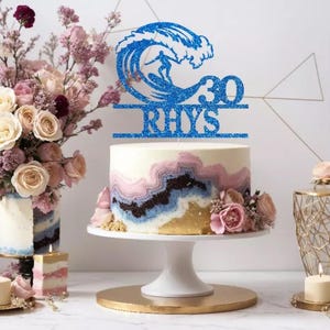 May include: Blue glitter cake topper featuring a surfer riding a wave, the number 30 and the name Rhys. The topper is on a white cake with pink, blue and gold frosting.