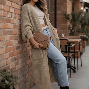 Könnte beinhalten: Eine Frau trägt einen langen, olivgrünen Strickcardigan, hellblaue Jeans und schwarze Stiefeletten. Sie hält eine braune, gesteppte Handtasche mit Kettenriemen. Im Hintergrund eine Backsteinmauer und Sitzgelegenheiten im Freien.