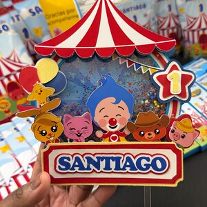 May include: A colorful circus-themed cake topper with a red and white striped tent design. The topper features cartoon characters, balloons, and the name "SANTIAGO" in blue letters. A star with the number "1" is also included.