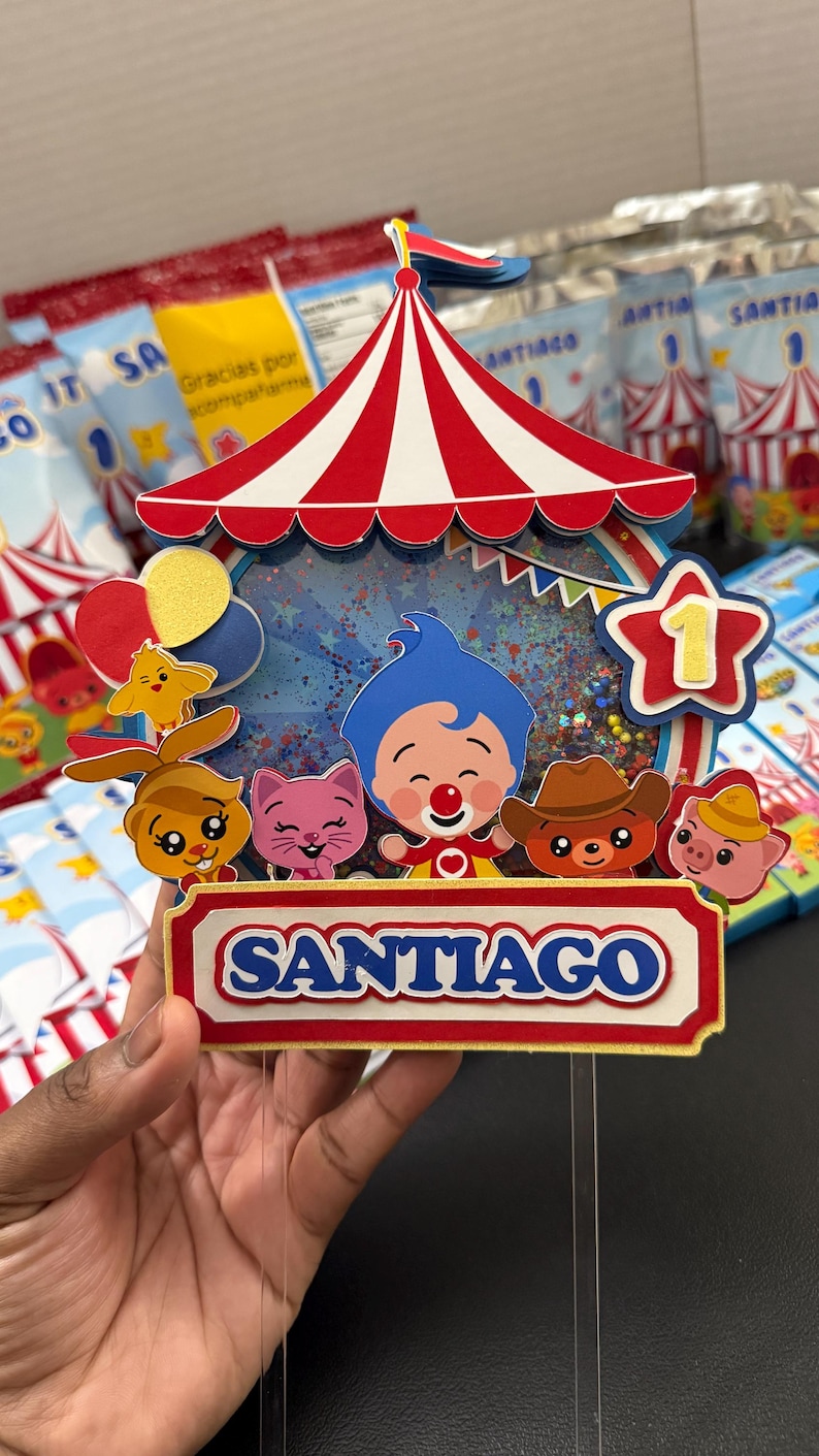 May include: A colorful circus-themed cake topper with a red and white striped tent design. The topper features cartoon characters, balloons, and the name "SANTIAGO" in blue letters. A star with the number "1" is also included.