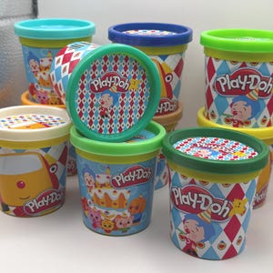 May include: A collection of Play-Doh containers with colorful lids and labels. The containers feature illustrations of cartoon characters and birthday themes. The lids have a diamond pattern. The Play-Doh logo is visible on the containers.