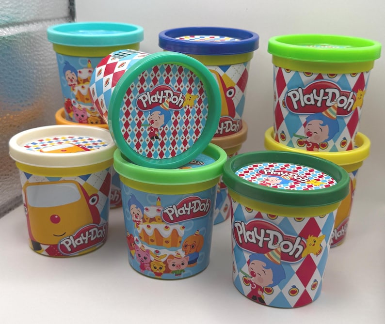 May include: A collection of Play-Doh containers with colorful lids and labels. The containers feature illustrations of cartoon characters and birthday themes. The lids have a diamond pattern. The Play-Doh logo is visible on the containers.