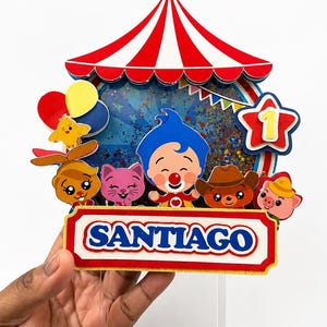 May include: A colorful circus-themed cake topper featuring a red and white striped tent, cartoon characters, and the name "SANTIAGO". The topper includes a star with the number "1" and balloons.