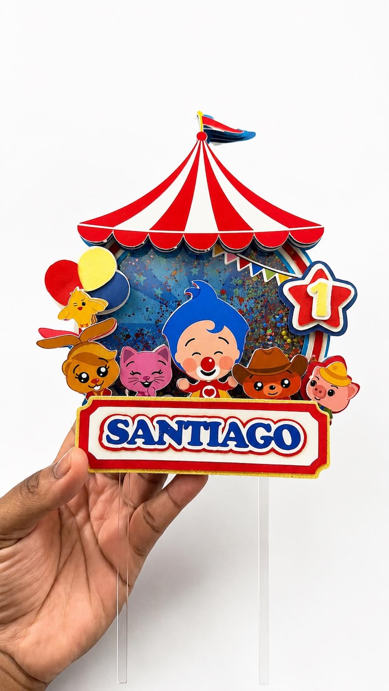 May include: A colorful circus-themed cake topper featuring a red and white striped tent, cartoon characters, and the name "SANTIAGO". The topper includes a star with the number "1" and balloons.