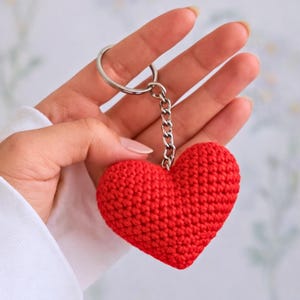 Puede incluir: Un llavero de corazón rojo tejido a mano. El dije en forma de corazón está unido a una cadena y un llavero de color plateado. El llavero es de color rojo vibrante y parece estar hecho de hilo suave.