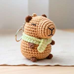 Puede incluir: Un llavero de capibara tejido a ganchillo en tonos marrones, con una bufanda verde claro. El peluche hecho a mano tiene un llavero plateado adjunto. El capibara tiene pequeñas orejas, pies y una cara cosida marrones.