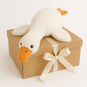 Peut inclure: Une peluche d'oie blanche avec un bec et des pieds orange est posée sur une boîte cadeau marron nouée avec un ruban et un nœud crème. L'oie a des yeux noirs et est faite d'une matière douce et moelleuse. La boîte est rectangulaire.