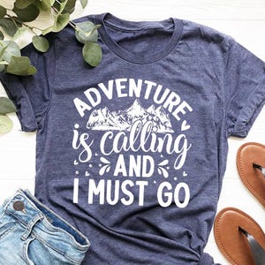 Może przedstawiać: Granatowa koszulka z białym napisem "ADVENTURE is calling AND I MUST GO". Koszulka jest połączona z dżinsowymi szortami i brązowymi sandałami. Gałązka zieleni znajduje się w lewym górnym rogu.