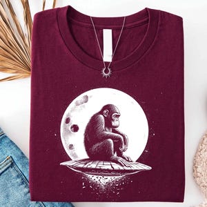 May include: A maroon t-shirt featuring a graphic of a chimpanzee sitting on a UFO in front of a full moon. A silver necklace with a sun pendant is draped over the shirt. The shirt is folded and displayed on a white surface.