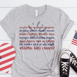 Puede incluir: Una camiseta gris con texto patriótico en rojo y azul, una decoración en forma de corazón y una pequeña bandera estadounidense. Zapatillas deportivas en blanco y negro completan el conjunto, ideal para el Día de la Independencia.