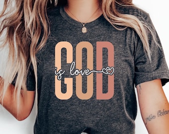 May include: A woman wearing a dark grey t-shirt with the text "GOD is love" in pink, orange, and yellow. The text is in a blocky font with a heart symbol at the end of the phrase.