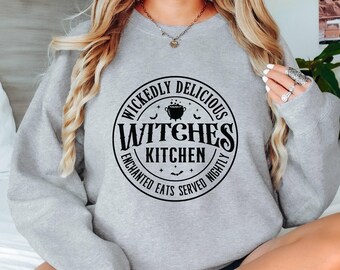 Könnte beinhalten: Grauer Pullover mit einem schwarzen Kreis-Motiv, auf dem "Wickedly Delicious Witches Kitchen Enchanted Eats Served Nightly" steht.