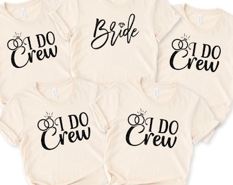Puede incluir: Cinco camisetas beis con texto negro. Una camiseta dice "Bride" con un diamante. Las otras cuatro camisetas dicen "I Do Crew" con un gráfico de anillo.