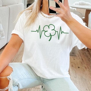 May include: A white t-shirt with a green four-leaf clover graphic and a heartbeat line. The shirt is being worn by a person with light brown hair and blue jeans.