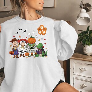 以下が含まれることがあります： ハロウィーンをテーマにしたグラフィックが特徴の白いスウェットシャツ。デザインには、ウッディ、ジェシー、バズ・ライトイヤー、カボチャ、恐竜に扮した漫画のキャラクターが含まれています。グラフィックには、コウモリ、幽霊、カボチャの風船も含まれています。