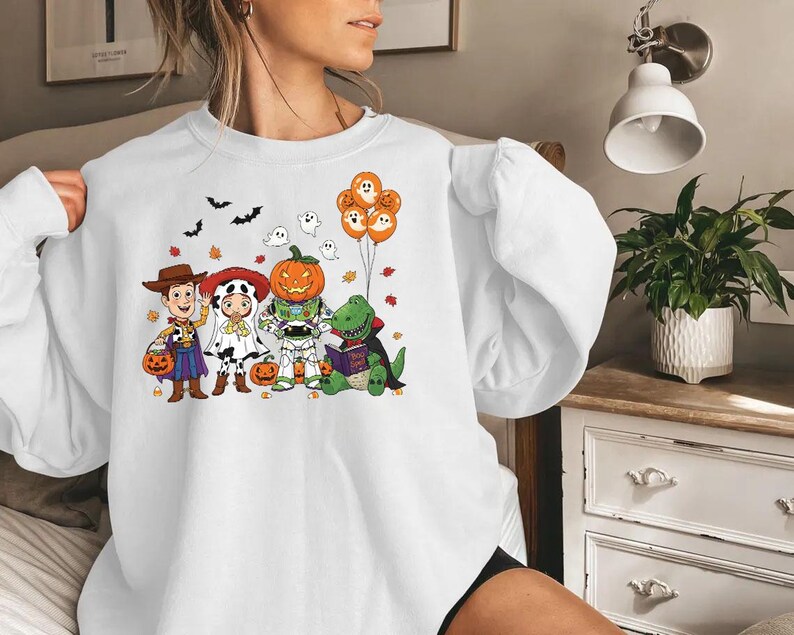 以下が含まれることがあります： ハロウィーンをテーマにしたグラフィックが特徴の白いスウェットシャツ。デザインには、ウッディ、ジェシー、バズ・ライトイヤー、カボチャ、恐竜に扮した漫画のキャラクターが含まれています。グラフィックには、コウモリ、幽霊、カボチャの風船も含まれています。
