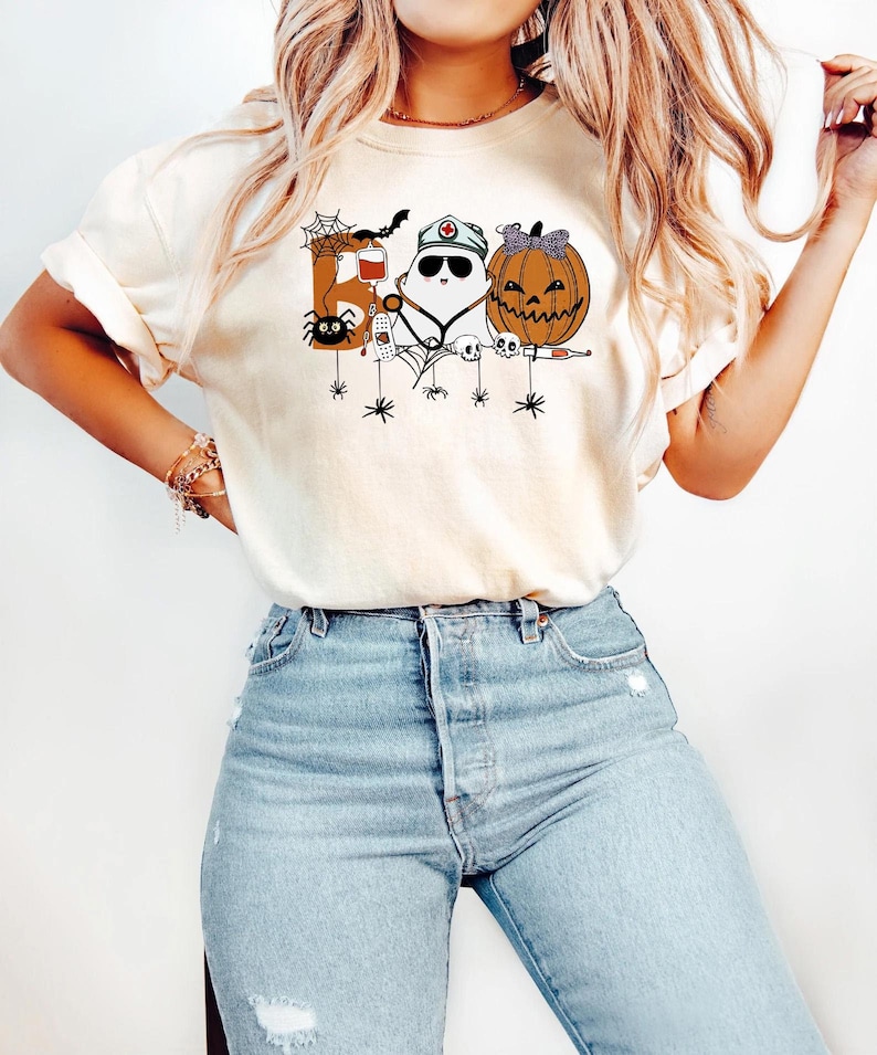 May include: Cream-colored Halloween-themed t-shirt featuring a graphic of a spider, a ghost wearing a nurse's cap, and a jack-o'-lantern. The design includes the word "Boo" and other Halloween elements. The shirt is paired with light blue jeans.