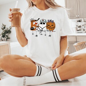 May include: White t-shirt featuring a Halloween-themed design with a letter, a ghost, and a jack-o'-lantern. The shirt has a crew neck and short sleeves. The design includes spiders and spiderwebs. The person is holding a glass of iced coffee.