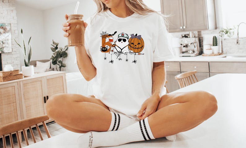 May include: White t-shirt featuring a Halloween-themed design with a letter, a ghost, and a jack-o'-lantern. The shirt has a crew neck and short sleeves. The design includes spiders and spiderwebs. The person is holding a glass of iced coffee.