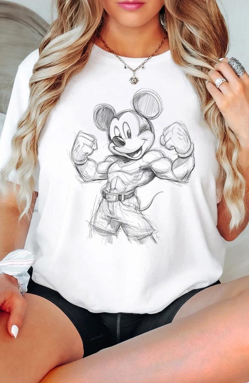 May include: A white short-sleeved t-shirt featuring a black and white sketch-style graphic of a muscular Mickey Mouse character flexing both arms. The character is depicted with defined chest and arm muscles, wearing shorts and a belt, rendered in a loose, hand-drawn pencil aesthetic on the front of the shirt.