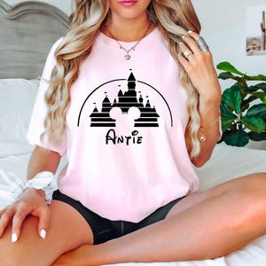 May include: A pink t-shirt featuring a black graphic of a castle and Mickey Mouse ears. The text "Auntie" is printed beneath the graphic.