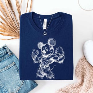 May include: A navy blue short-sleeved t-shirt featuring a white line-art sketch of a muscular cartoon mouse character flexing its arms. The shirt has a crew neckline and a white brand label at the inner collar, displayed with a silver sun pendant.